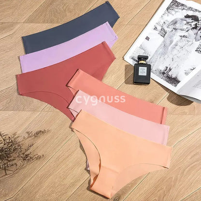 Imagem de calcinhas sem costura da Cygnuss dispostas sobre um fundo de madeira, apresentando tons suaves e quentes, como lilás, cinza, terracota, rosa e pêssego. Ao lado, um frasco de perfume e uma revista complementam a composição sofisticada.