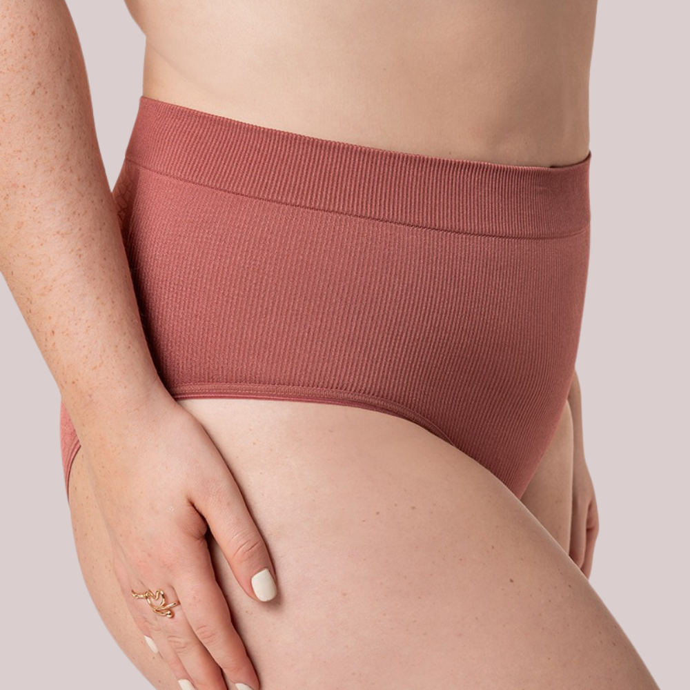 Close-up lateral de uma mulher usando uma calcinha modeladora de cintura alta na cor telha. O tecido canelado sem costuras oferece ajuste confortável e efeito modelador suave, realçando a silhueta. A cintura elástica molda o corpo sem apertar, proporcionando sustentação e bem-estar. O logotipo da Cygnuss & Lupo está sobreposto na parte inferior da peça. Fundo nude rosado. 