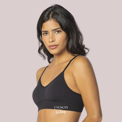 Mulher de pele morena e cabelos ondulados usando um sutiã preto sem costuras, com alças finas e modelagem anatômica. O tecido respirável e macio proporciona conforto e ajuste perfeito ao corpo. O logotipo da Cygnuss & Lupo está sobreposto na parte inferior da peça. Fundo nude rosado.
